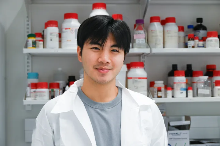 Sean stands in his white lab coat in front of a wall of medicine.