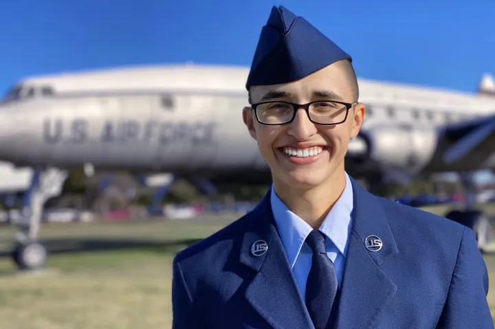 Jacob Flores smiles at the camera wearing his full Air Force uniform, with an Air Force plane behind him in the distance.