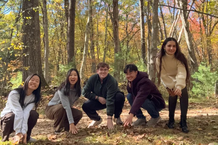 A group of students crouch and smile while touching the ground of Harvard Forest.
