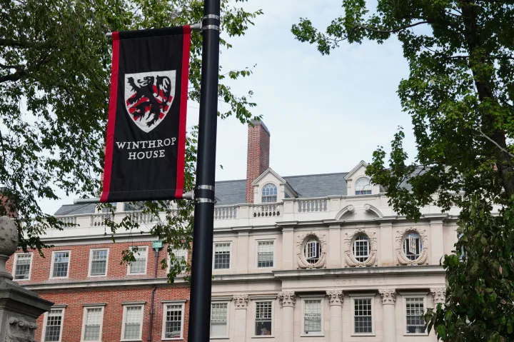 A banner with a crest and "Winthrop House" hangs on a pole in front of a historic brick and stone building, surrounded by trees.