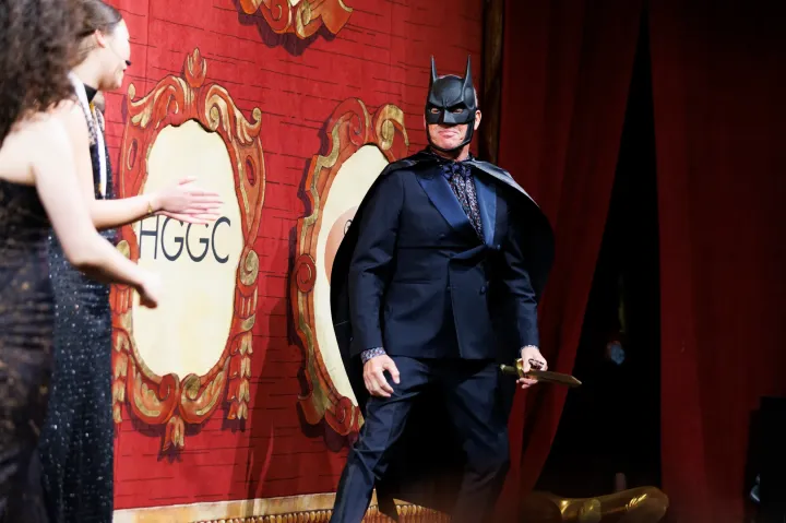 Person in a Batman mask and cape stands on stage in a suit, with a red curtain and another person clapping.