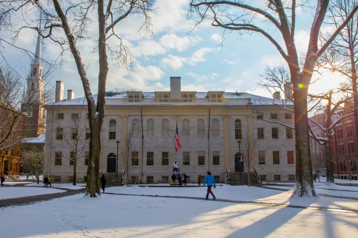 Historic campus building with an American flag and statue in front, surrounded by snow and tall trees with people walking.