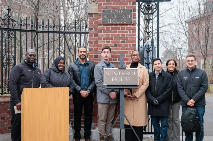 Group of people stand smiling by the Winthrop House gate and sign during an outdoor ceremony.