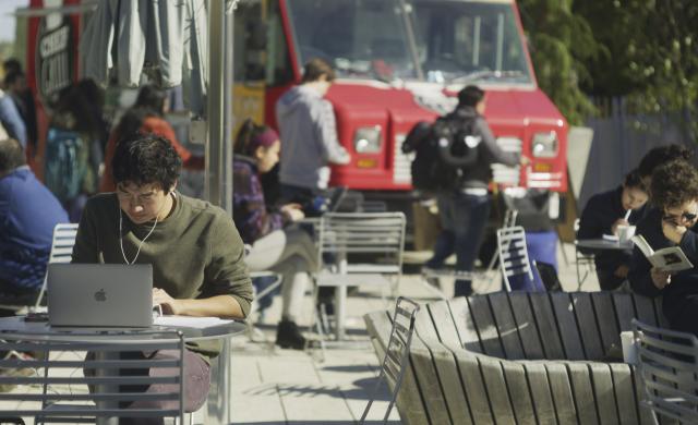 A student in a green sweater sits at a laptop at an outside table. Students in the background are getting lunch from a foodtruck.