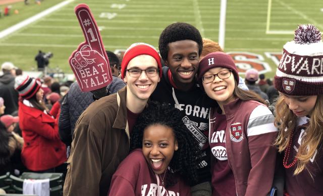 Four students cheering in the stands at a Harvard football game