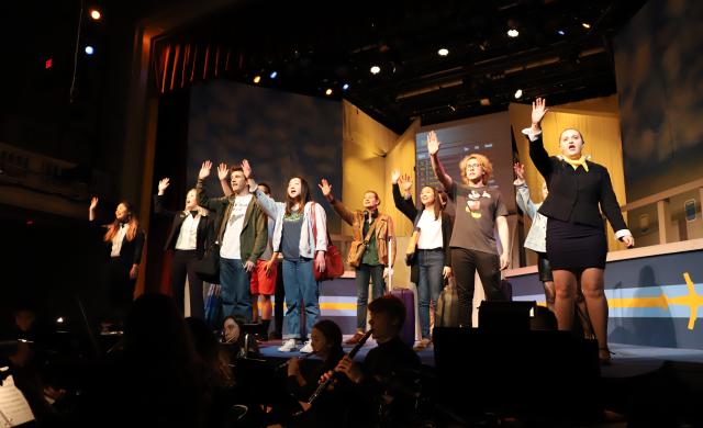 Actors on stage raising their hands singing, with musicians below