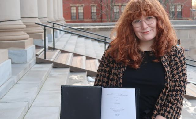 Allison standing on the steps of a large library holding a black thesis binder opened to the title page of her thesis.
