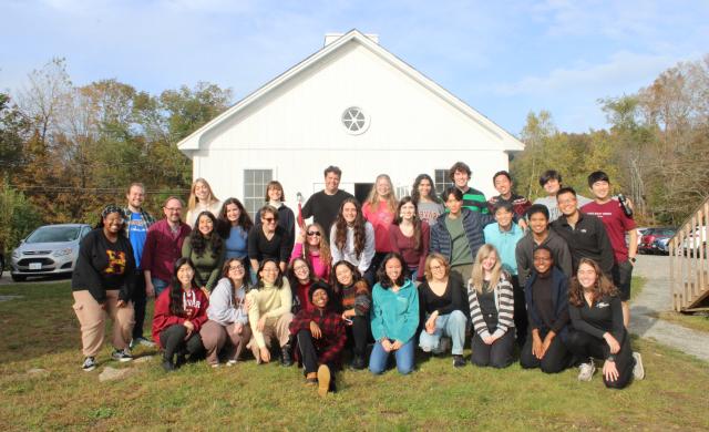 Picture of the Harvard-Radcliffe Collegium Musicum standing in front of a white cabin under blue skies on a green hill.