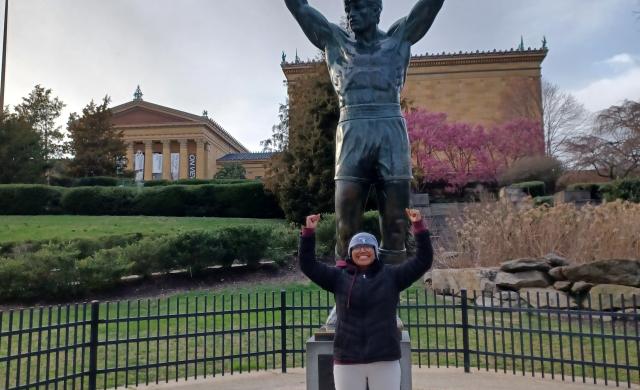 Merlin standing in front of Rocky Statue with fists in the area in front of the steps where the Rocky movie scene was shot