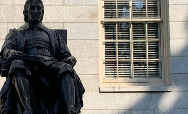 This is a picture of the top of the John Harvard Statue, which shows only the shape of the statue itself, and not the base with it's description. The statue shows a man posing in a sitting, very academic looking, position. 