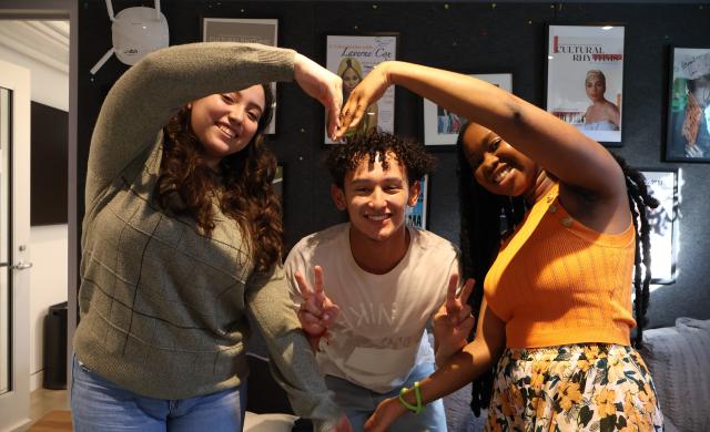 This is a picture of three smiling individuals posing together by forming a heart shape with their arms. The person on the left wears a green sweater and has long, wavy hair. The person in the middle, wearing a white T-shirt, flashes peace signs with both hands, while the person on the right, dressed in a bright orange top and floral skirt, completes the heart shape with a big smile. The background features framed posters and a cozy room ambiance.