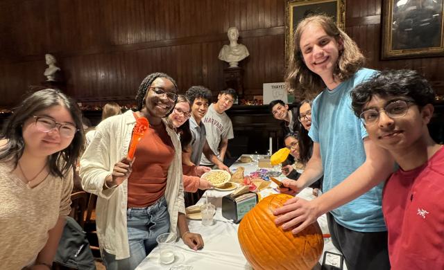 Students holding pumpkin-carving tools stand around a table in Annenberg Hall. On the table sits a pumpkin and several pieces of candy.