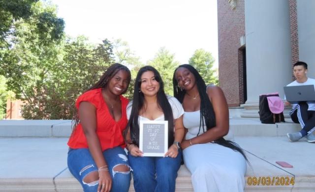 My friends and I sitting on the steps of Widener Library, holding a sign that says "Happy First day of 14th Grade"."