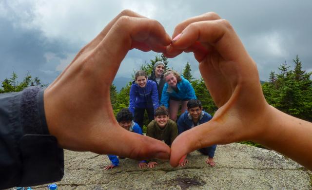 A group of six first-year students on a FOP trip form a human pyramid atop a mountain summit, framed by the hands of their FOP leaders, which form a heart shape.