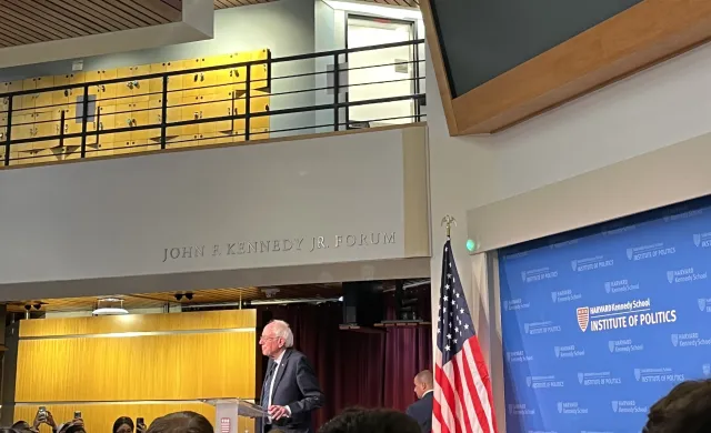 A picture of the main lobby of the Kennedy School building. It is decorated with an American flag and full of students listening to Bernie Sanders speak.