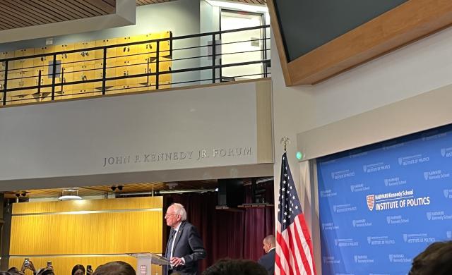 A picture of the main lobby of the Kennedy School building. It is decorated with an American flag and full of students listening to Bernie Sanders speak.