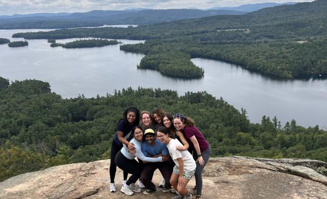 Lizbeth and seven peers posing on top of a mountain in New Hampshire, which a large lake in the background.