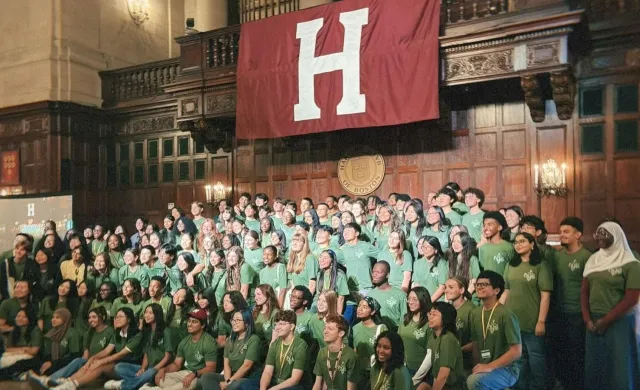 A large group of diverse students wearing matching green t-shirts posing for a group photo inside a grand, wood-paneled hall. They are gathered beneath a large crimson banner displaying a white letter "H" and a circular Harvard Club of Boston emblem.