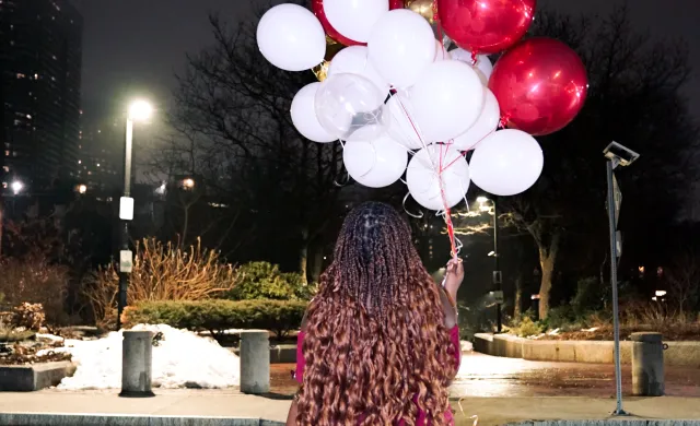 Photo of girl in red dress holding red and white balloons while walking across the street
