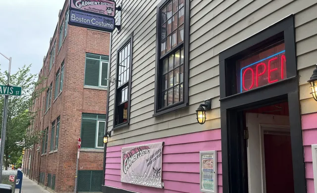 Gray and pink building with lamp lights and a neon "OPEN" sign above the door. Two signs protrude from the building, saying "The Garment District" and "Boston Costume" 