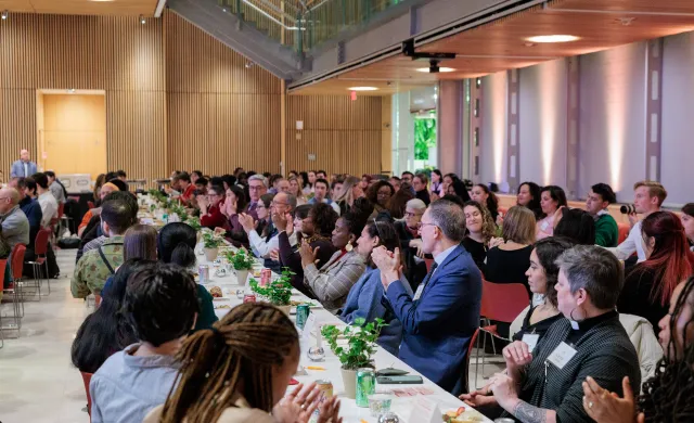 Many students, faculty, and staff sit at several long tables, eating and talking