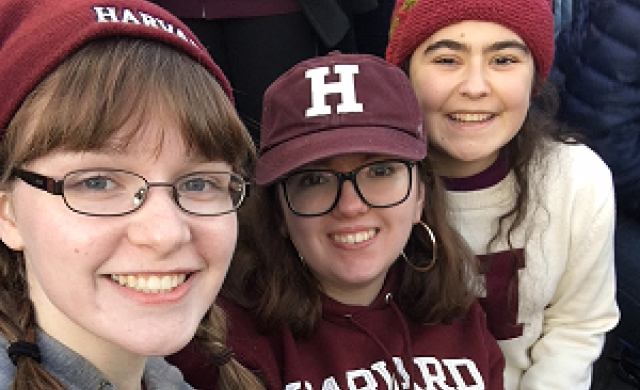 Three girls in Harvard gear take a selfie in the bleachers of a football stadium.