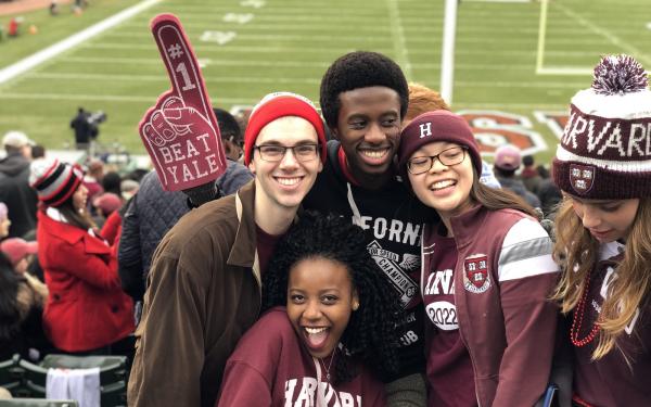 Four students cheering in the stands at a Harvard football game