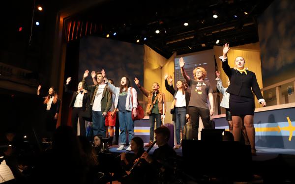 Actors on stage raising their hands singing, with musicians below