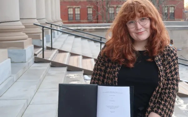 Allison standing on the steps of a large library holding a black thesis binder opened to the title page of her thesis.