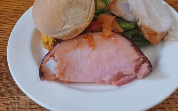 White plate piled with a slice of ham, turkey, a bread roll, green beans, and sweet yams.