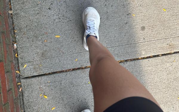 A girl&#039;s tan leg wearing black running shorts and white running sneakers, mid stride on a concrete sidewalk.