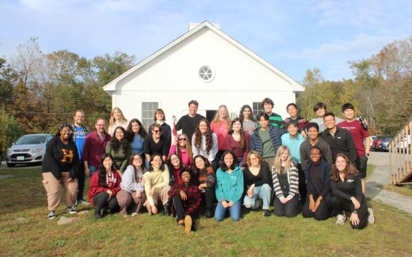 Picture of the Harvard-Radcliffe Collegium Musicum standing in front of a white cabin under blue skies on a green hill.