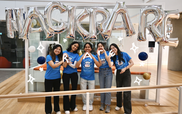 A group of five students wearing blue shirts in front of silver balloons that read "HACK RARE." They are making symbols with their hands to spell out "RARE."