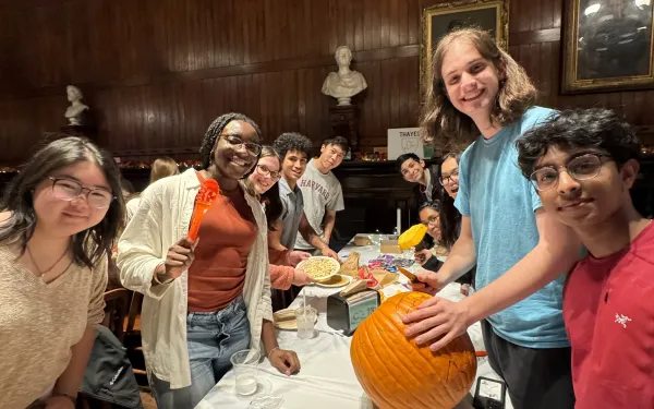 Students holding pumpkin-carving tools stand around a table in Annenberg Hall. On the table sits a pumpkin and several pieces of candy.