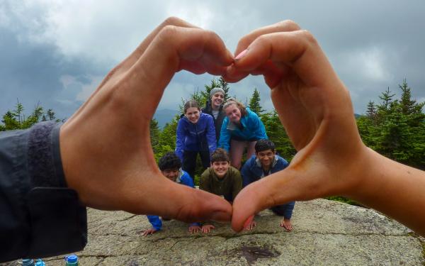 A group of six first-year students on a FOP trip form a human pyramid atop a mountain summit, framed by the hands of their FOP leaders, which form a heart shape.