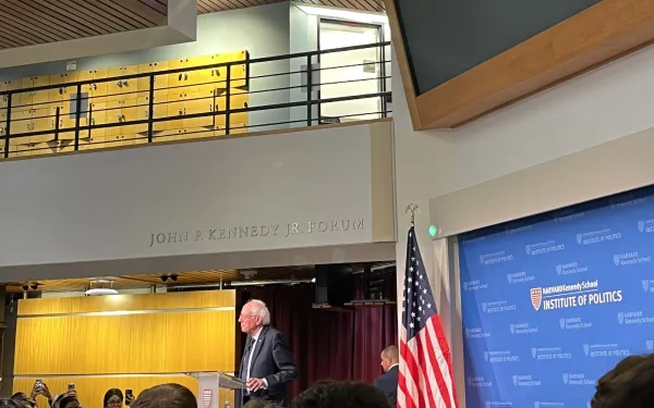 A picture of the main lobby of the Kennedy School building. It is decorated with an American flag and full of students listening to Bernie Sanders speak.