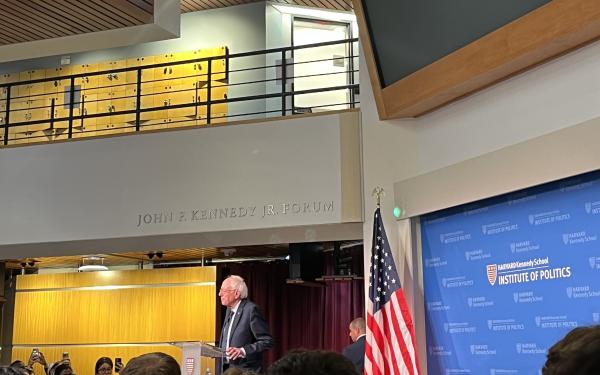 A picture of the main lobby of the Kennedy School building. It is decorated with an American flag and full of students listening to Bernie Sanders speak.