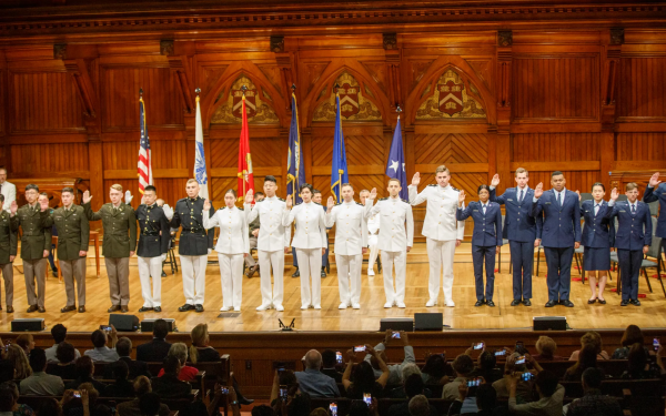 A group of military cadets in uniform stand on stage with their right hands raised during a ceremony.