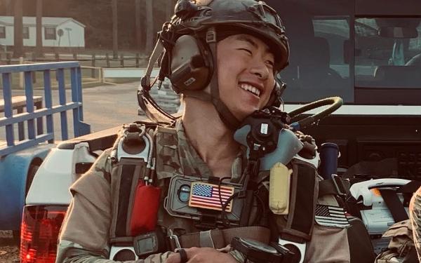 A smiling soldier in camouflage gear and helmet stands in front of a vehicle.