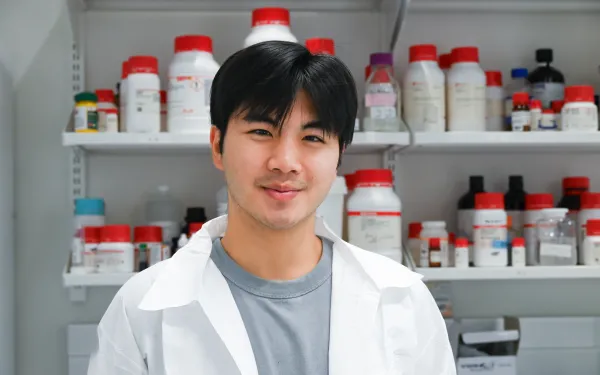 Sean stands in his white lab coat in front of a wall of medicine.