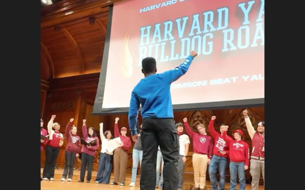 Person leads a group on stage with raised fists in front of a large "Harvard Yale Bulldog Roast" screen.