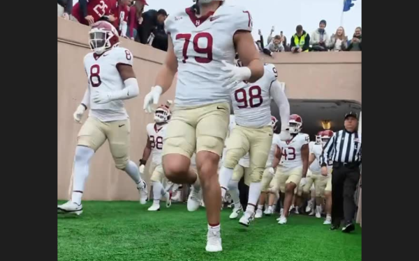 Harvard football players in white jerseys run out of a tunnel onto the field.