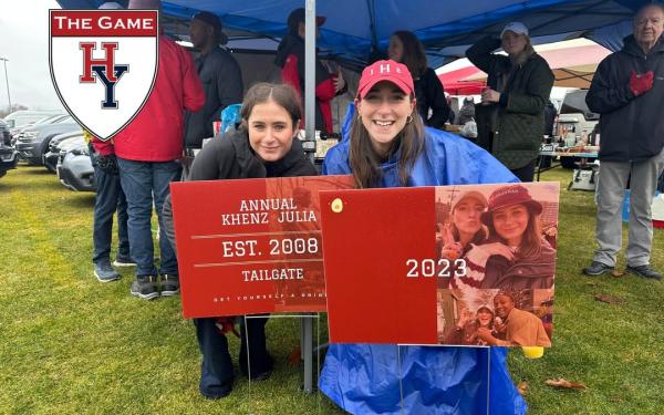 Two women pose at a tailgate, holding signs for "The Game" with Harvard-Yale logos.