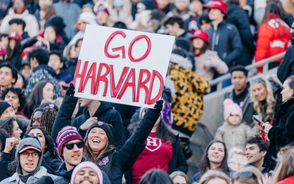 Person in a crowd holding a large "GO HARVARD" sign at a sports event.