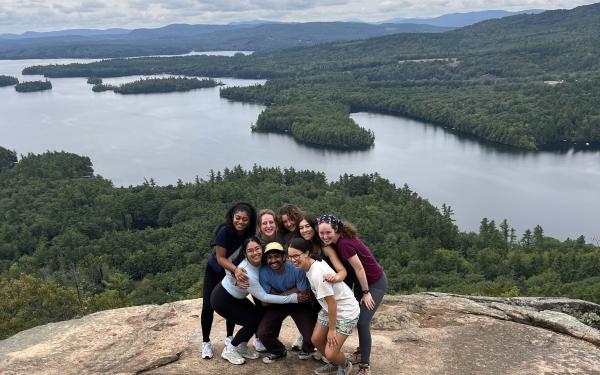 Lizbeth and seven peers posing on top of a mountain in New Hampshire, which a large lake in the background.