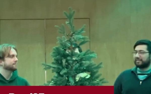 Two students stand onstage on either side of a small evergreen tree