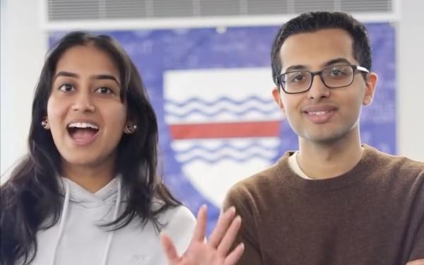Two students stand indoors in front of a blue shield banner.