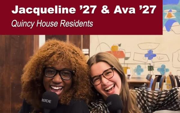 Two Quincy House juniors, Jacqueline and Ava, sit together smiling and holding microphones in a dining hall.