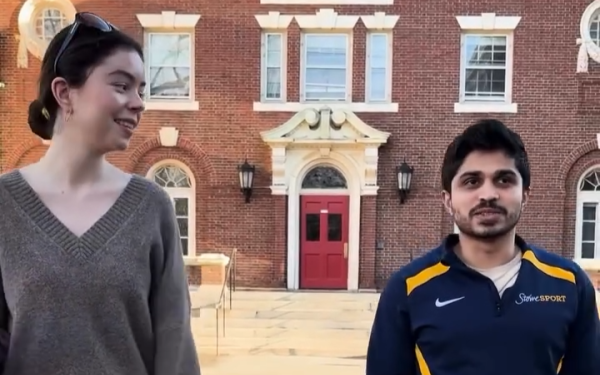 Two students talk outside a red-brick campus building with a red door and arched windows.