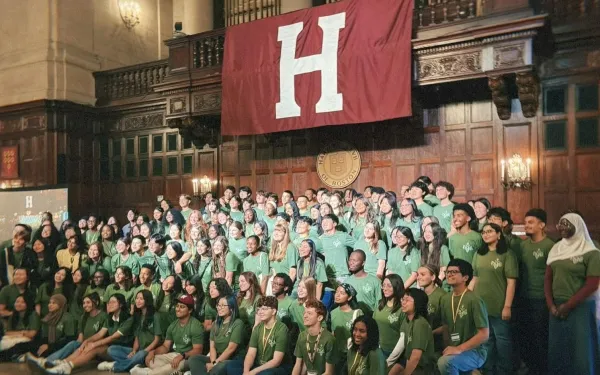 A large group of diverse students wearing matching green t-shirts posing for a group photo inside a grand, wood-paneled hall. They are gathered beneath a large crimson banner displaying a white letter "H" and a circular Harvard Club of Boston emblem.
